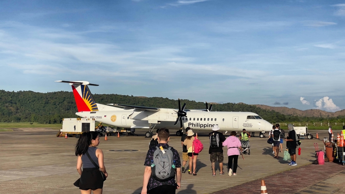 空港の駐機場にて、フィリピン航空の白い機体に向かって歩いていく乗客たちの後ろ姿。リュックを背負った人々が、晴天の下、山々に囲まれた広々とした滑走路を機体へと進んでいる。