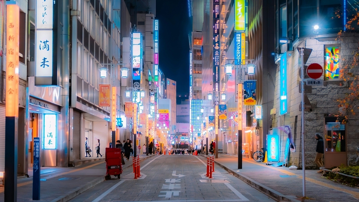 夜の新宿西口商店街。立ち並ぶビルに青やオレンジの鮮やかなネオン看板が点灯し、通りを歩く人々の姿が見える都会の夜景です。