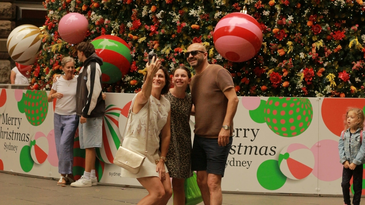 「Merry Christmas Sydney」と書かれたパネルの前で、大きなオーナメントが飾られたツリーを背景に自撮りをする家族