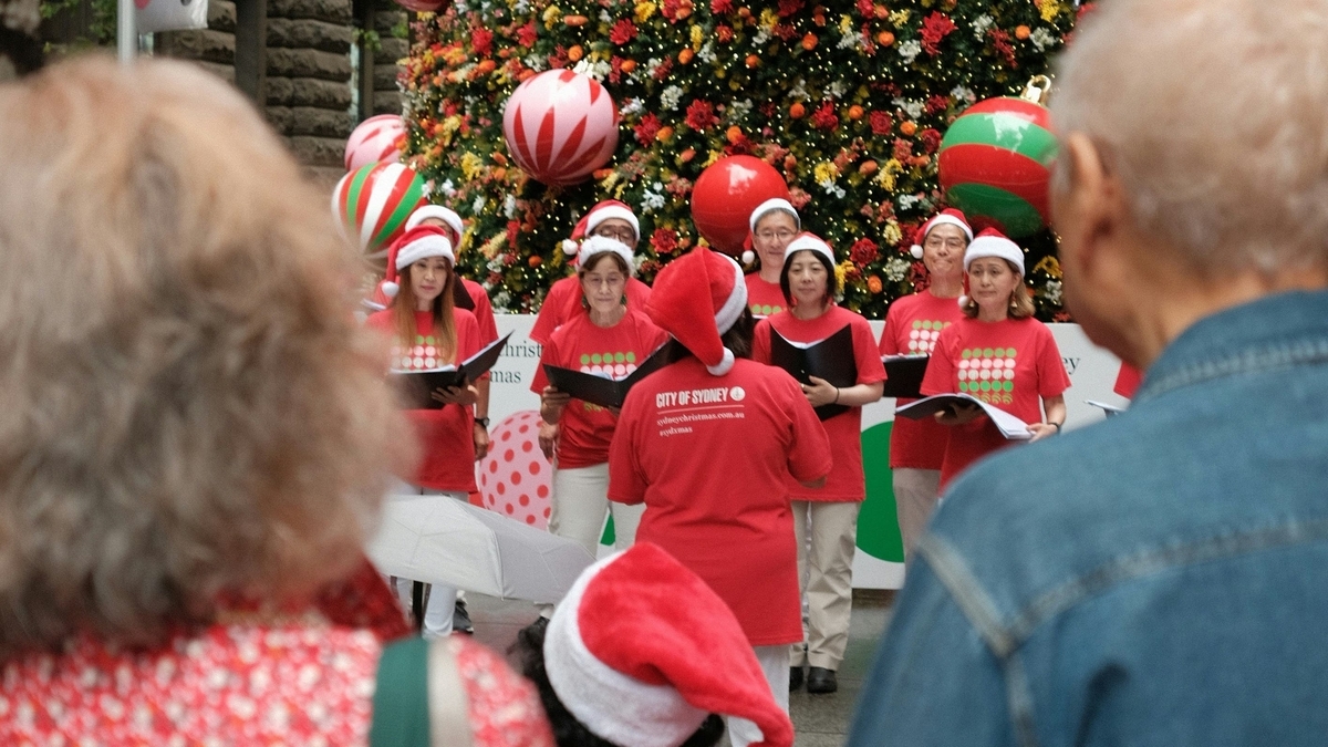 サンタクロースの帽子と赤いTシャツを揃えて着用し、大きなクリスマスツリーの前で合唱する聖歌隊の男女