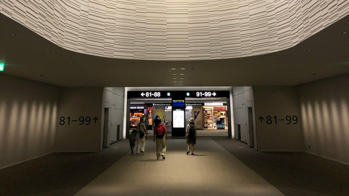 空港内の通路で「81-99」と書かれた案内表示に従い、搭乗ゲート方面へ向かって歩く旅行者たち