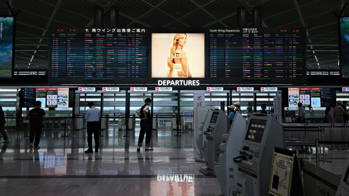 空港の出発ロビーに設置された、多くの便名が並ぶ電光掲示板と自動チェックイン機が並ぶ様子