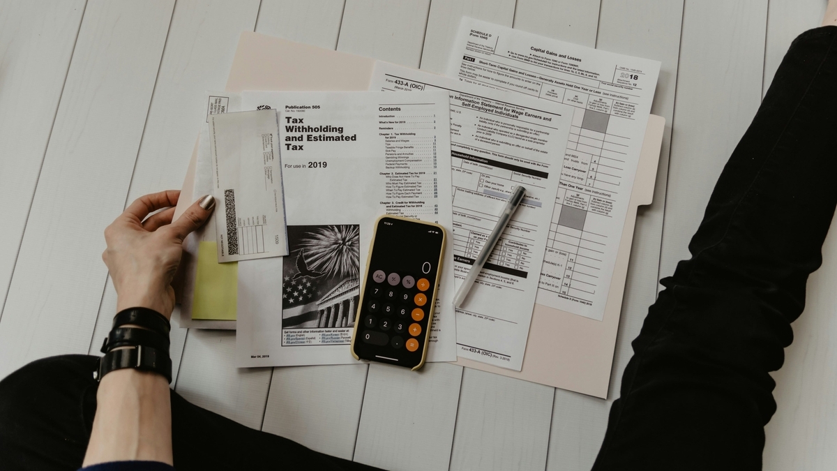 フローリングに座る人の膝の上で、2019年の納税に関する書類や封筒が広げられ、中央にスマートフォンの計算機画面が見える。