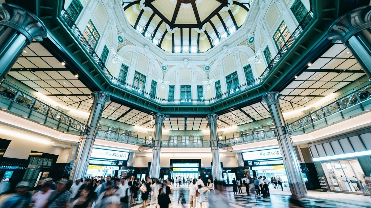 東京駅、高い天井のドーム構造を持つ巨大な駅舎のホールで、多くの人々が行き交い、周囲を巨大な柱と装飾豊かな壁面が囲む広大な空間