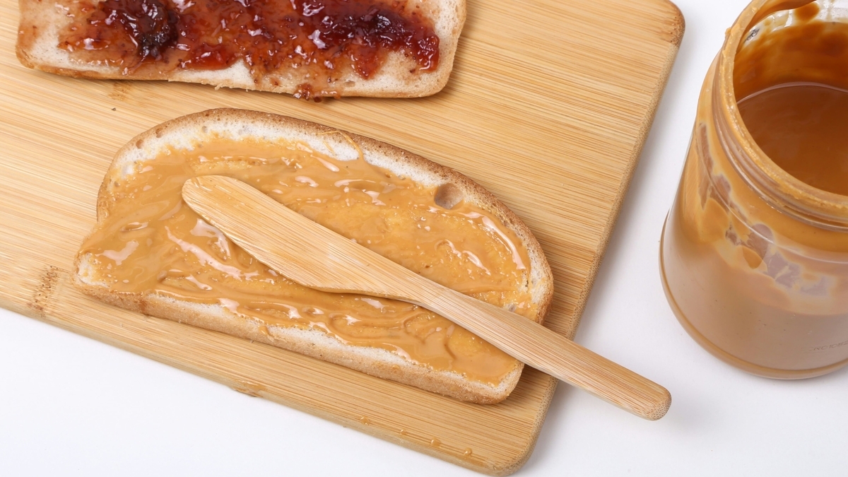 木のまな板の上で、一切れの食パンに木製のヘラを使ってなめらかなピーナッツバターを塗る様子。奥にはジャムを塗ったパンとピーナッツバターの瓶が置かれています。