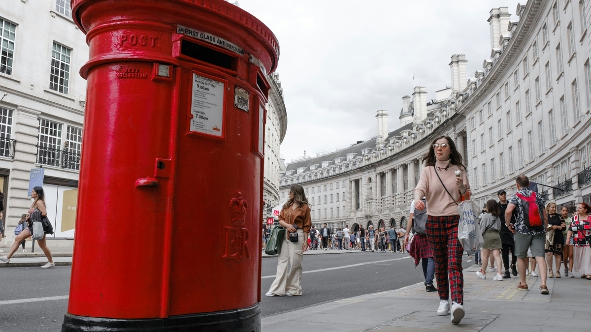 ロンドンの街角に設置された、重厚感のある赤い円筒形のポスト。背景には緩やかにカーブした白いクラシックな建物が並び、歩道を多くの人々が行き交う日常の風景が広がっています。