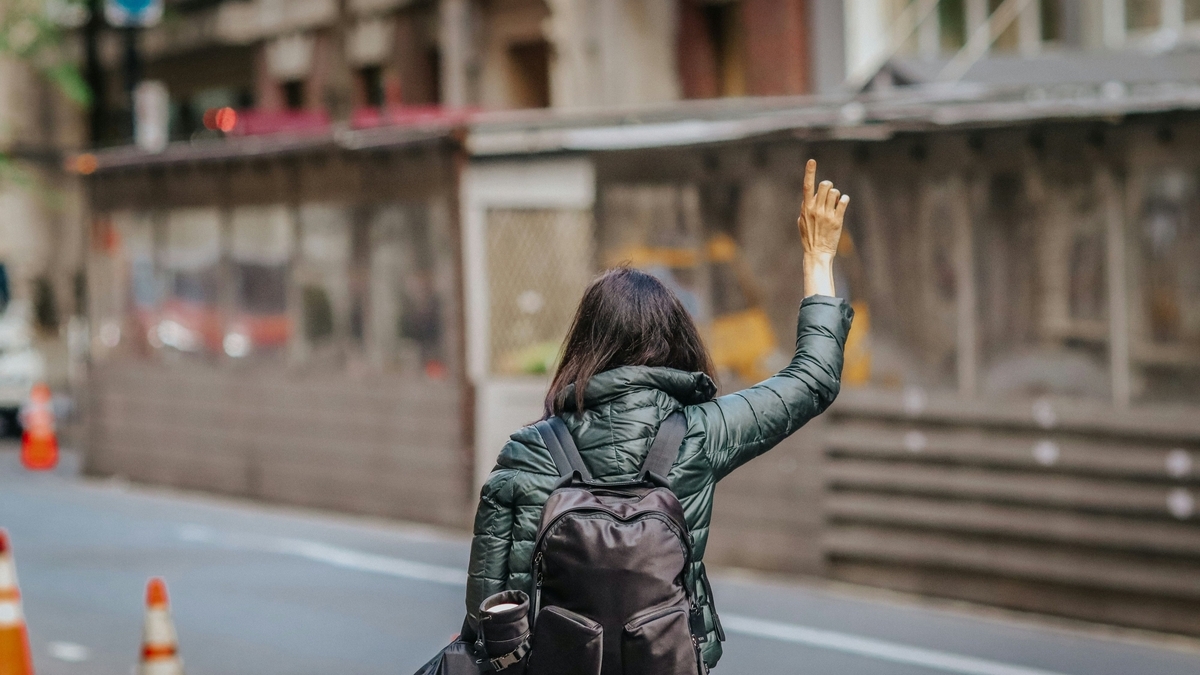 ウンタウンの歩道に立ち、右手を高く挙げてタクシーを呼ぶ女性の後ろ姿。緑のダウンジャケットに黒いバックパックを背負い、通り過ぎる車に向けて指を一本立てて合図を送っています。