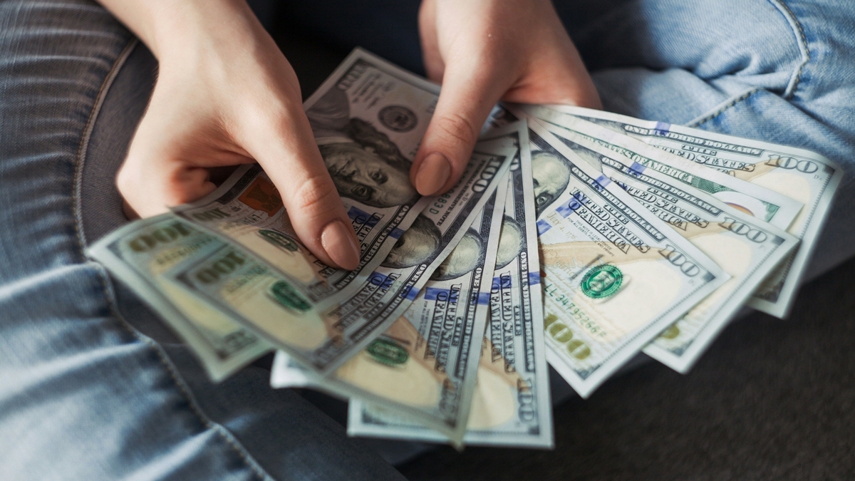 ジーンズを履いた人の手元で、扇状に広げられた複数枚の100米ドル紙幣