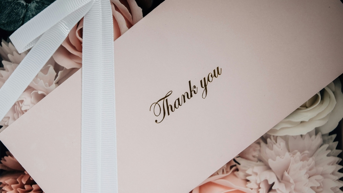 ピンク色の花々に囲まれた、「Thank you」という金色の文字が印字されたメッセージカード。白いリボンが斜めにかけられている