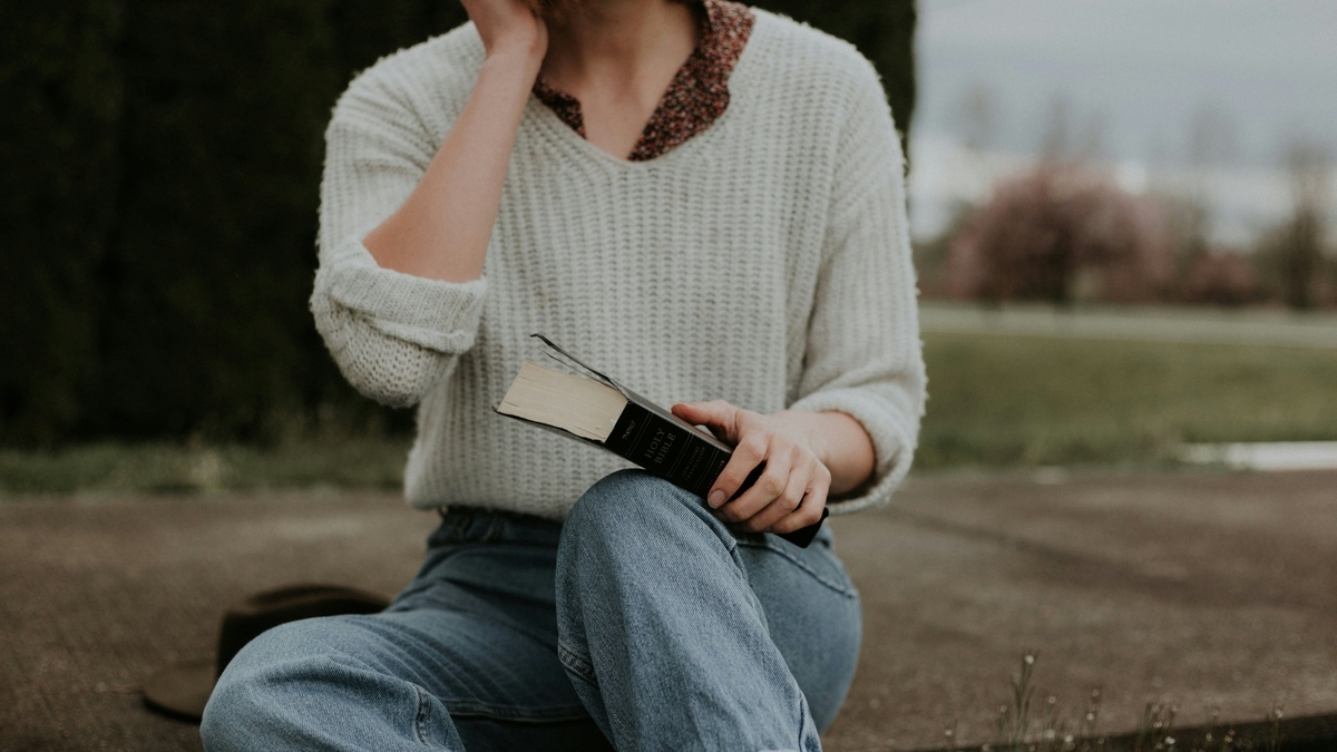 白いニットとジーンズ姿の人物が屋外の段差に座り、膝の上に黒い表紙の「HOLY BIBLE」と書かれた本を置いて手に持っている。