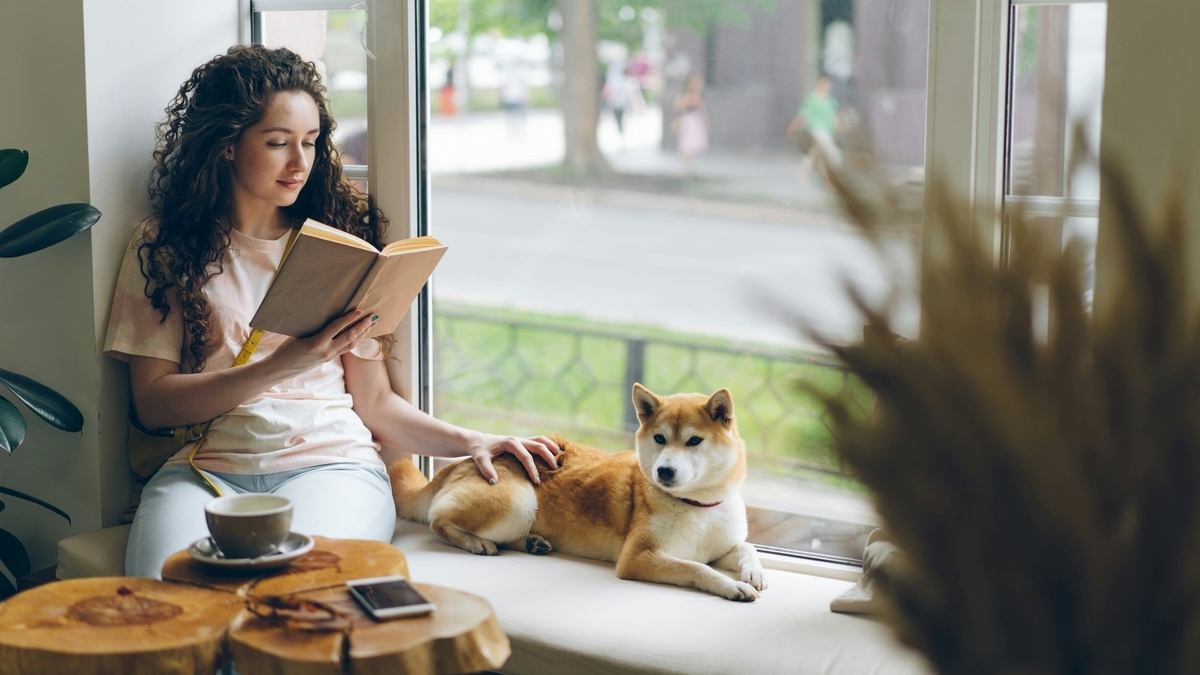 明るい窓際で読書を楽しむロングヘアの女性と、その傍らでくつろぐ柴犬。女性は片手で犬を撫でており、手前の切り株風のテーブルにはコーヒーカップとスマートフォンが置かれています。