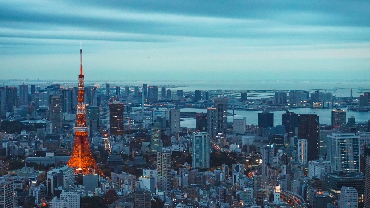 薄暗い夕景の中にオレンジ色にライトアップされた東京タワーが輝く都会の夜景。遠くには東京湾とベイエリアの明かりが広がり、重なり合うビル群が地平線まで続いています