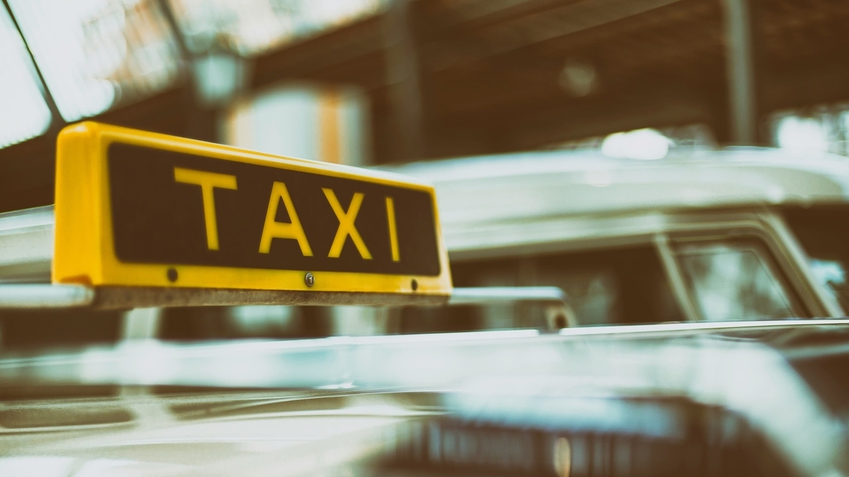 タクシーの車体屋根に取り付けられた、黄色いフレームに黒地の「TAXI」という文字が書かれた表示灯のクローズアップ。背景には駅のホームのような構造物と別の車両がぼけて写っている様子