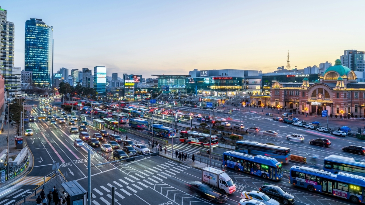 夕暮れ時のソウル駅周辺の街並み。歴史的なドーム屋根の旧駅舎と近代的な駅ビルが並び、手前の広い多車線道路には青い路線バスや乗用車が数多く行き交う、活気ある都市の景観