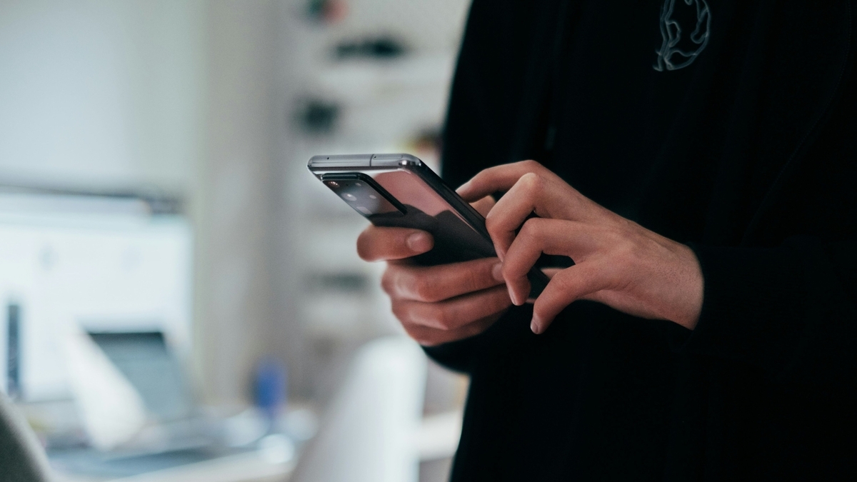 黒い服を着た人物が、両手でスマートフォンを操作している手元のアップ。スマートフォンの背面には複数のカメラレンズが見え、背景はデスクやモニターがぼやけて写っている