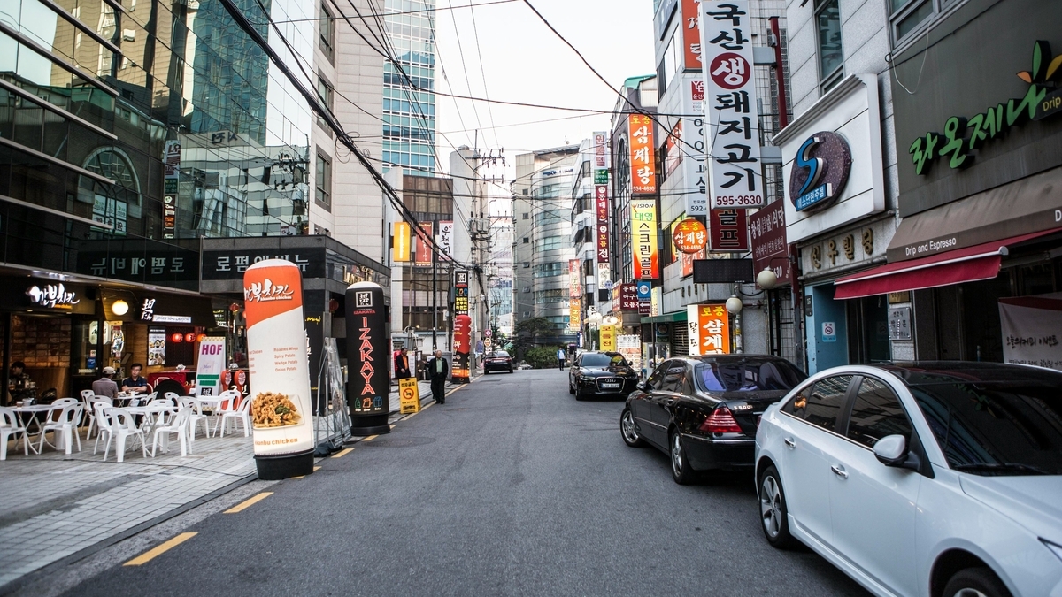 ハングルの看板がひしめく韓国の繁華街の路地。左手にはテラス席のある飲食店があり、路上には複数の乗用車が停車している。奥には高層ビルが見え、電線が空に張り巡らされている