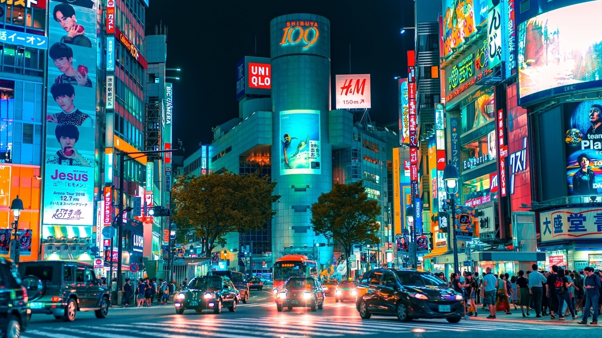 夜の渋谷109周辺の交差点。ネオン看板やビルボードが街を明るく照らし、道路にはヘッドライトを点灯させた車やバスが並んでいます。歩道には多くの人々が行き交う、活気あふれる都市の夜景です