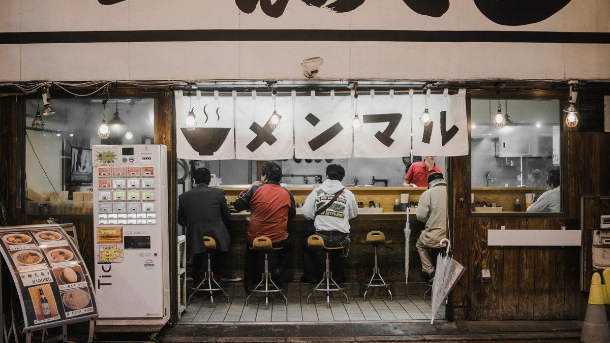 「メンマル」と書かれた白い暖簾がかかる、夜のラーメン店の外観。カウンター席に座り食事をする背広姿などの客や、券売機、店内の厨房で働く店員の姿が、開かれた入り口から見えています