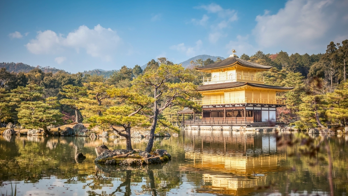鏡のような池のほとりに建つ、金箔で覆われた三層構造の舎利殿（金閣）。背後には緑豊かな山々が連なり、青空の下で黄金色の建物が水面に鮮明に反射している美しい風景です
