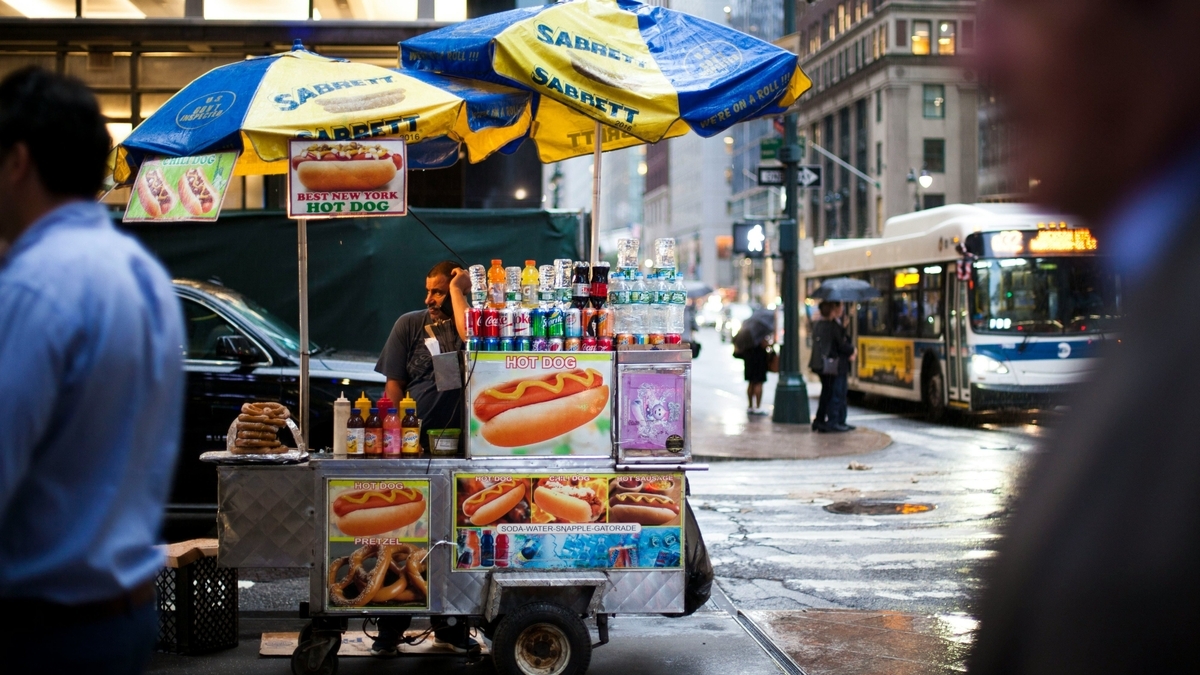 青と黄色のパラソルを立てたホットドッグの屋台が雨上がりの路上で営業し、背後を白いバスが通り過ぎる様子