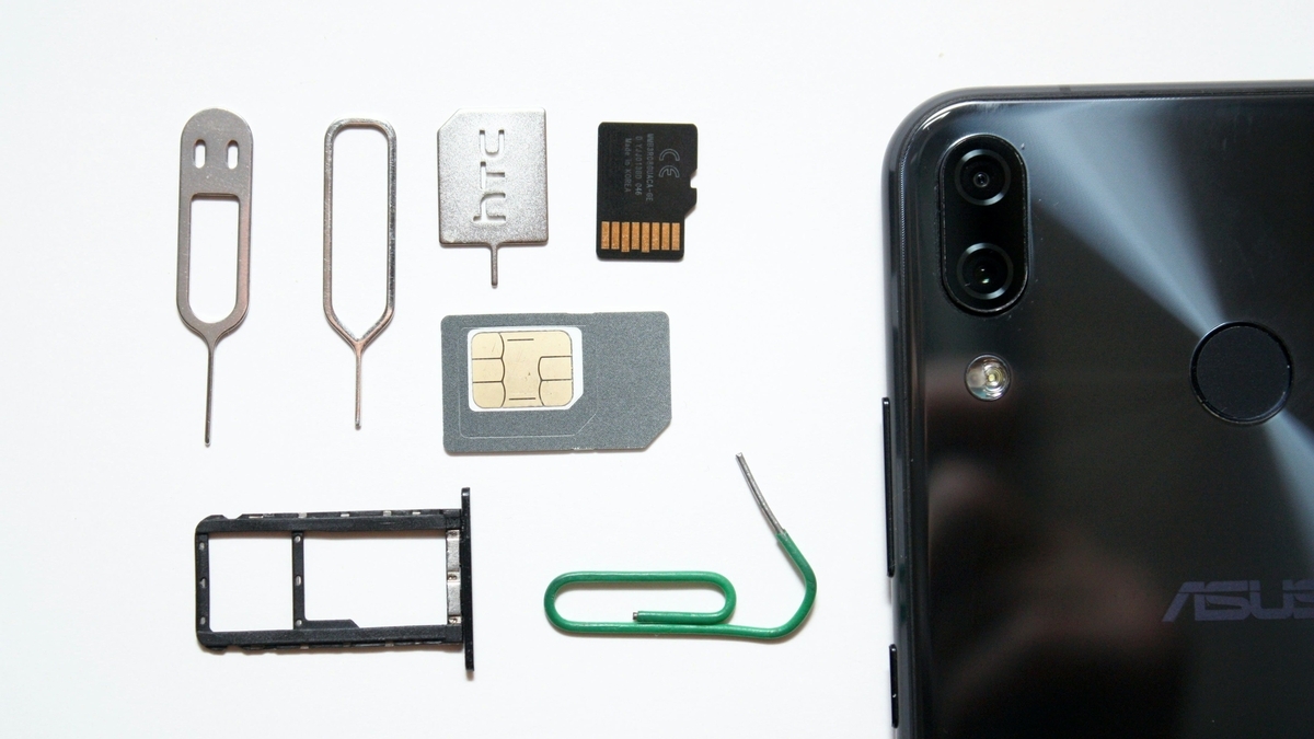 白背景の上に、黒いスマートフォンの背面と、その横に並べられたSIMカード、microSDカード、SIM取り出し用ピン、SIMトレイ、緑色のクリップといった小さな電子部品や周辺小物
