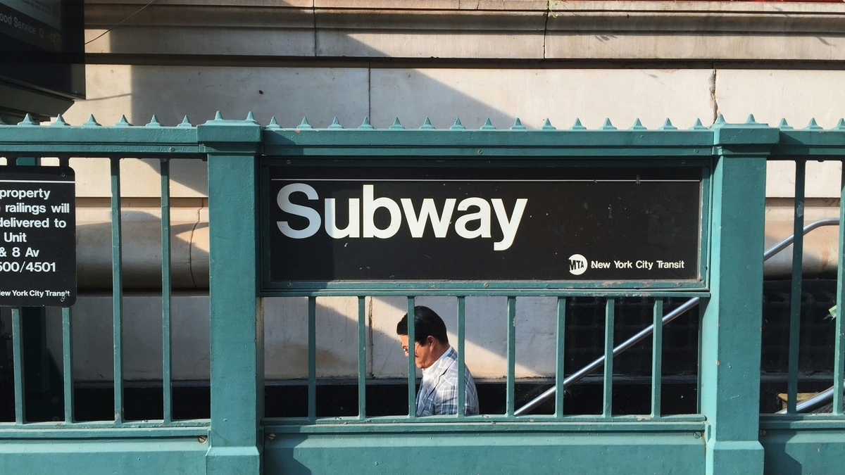 緑色の鉄柵に掲げられた「Subway」の看板と、その奥にある地下鉄の入り口へと続く階段を降りていく、チェック柄のジャケットを着た男性の横顔を捉えたニューヨークの街角