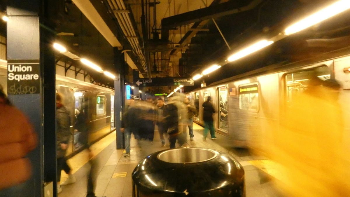 ニューヨークのユニオンスクエア駅のホーム。柱に「Union Square」と書かれた看板があり、左右に停車する地下鉄車両と、移動する人々が動体検知でブレて写っている活気ある風景