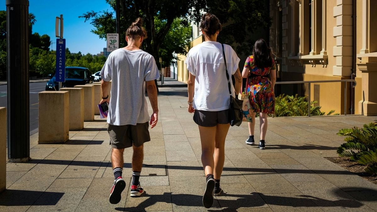 晴天の歩道を歩く男女の後ろ姿。男性はグレーのTシャツとハーフパンツ、女性は白いTシャツと黒のショートパンツを着用し、奥にはカラフルなワンピースを着た女性や街路樹、標識、建物が見える街並み
