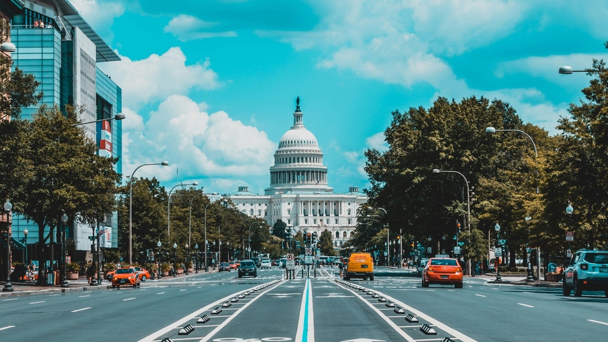 青空と白い雲を背景に、広い道路の先に白いドームを持つアメリカ合衆国議会議事堂が、緑の木々に挟まれて立っている