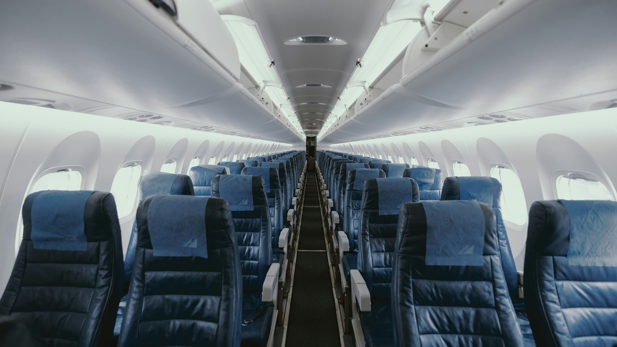 青いヘッドレストカバーがついた座席が通路の両側に整然と並ぶ、誰もいない旅客機の機内。天井の照明が点灯し、奥まで続く通路が見える