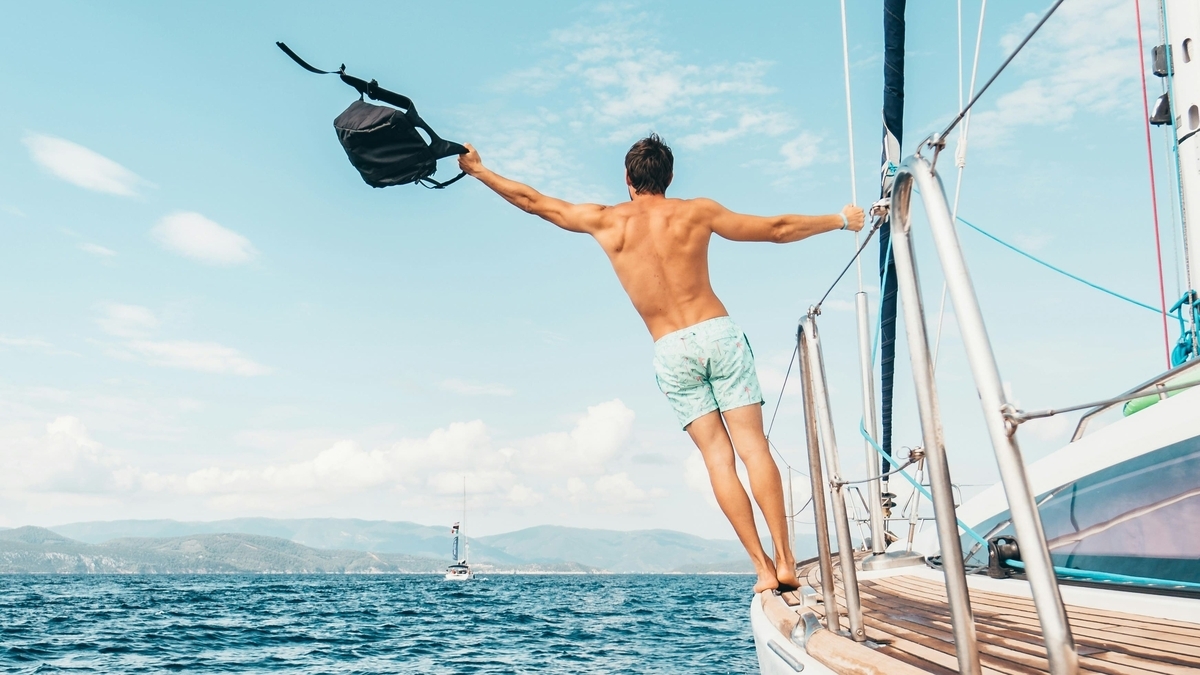 青く澄んだ海の上を走行する白いヨットの船尾で、水色の水着を着た男性が黒いリュックを片手で掲げながら、両腕を大きく広げてバランスを取っている後ろ姿