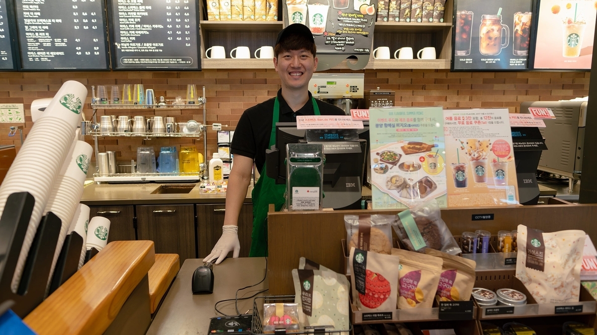 スターバックスのカウンター内で、緑色のエプロンを着用した男性店員が笑顔で立っている。背後の棚にはメニュー表やカップが並び、手前には個包装されたフード類が配置されている