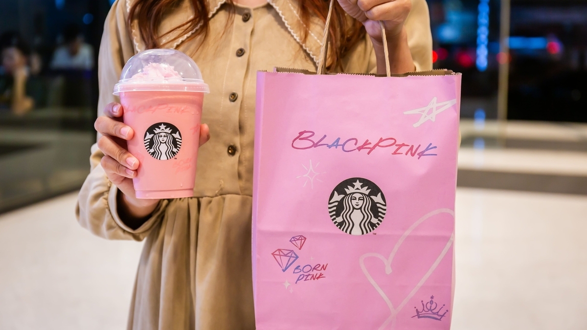 ベージュのワンピースを着た女性が、スターバックスのロゴが入ったピンク色のフラペチーノと、BLACKPINKのロゴが描かれたピンク色の紙袋を両手に持っている様子