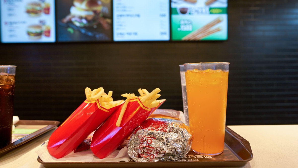 トレーの上に置かれた、マクドナルドのポテト2個と銀紙に包まれたハンバーガー、オレンジ色の炭酸飲料。背景にはメニューボードが見える