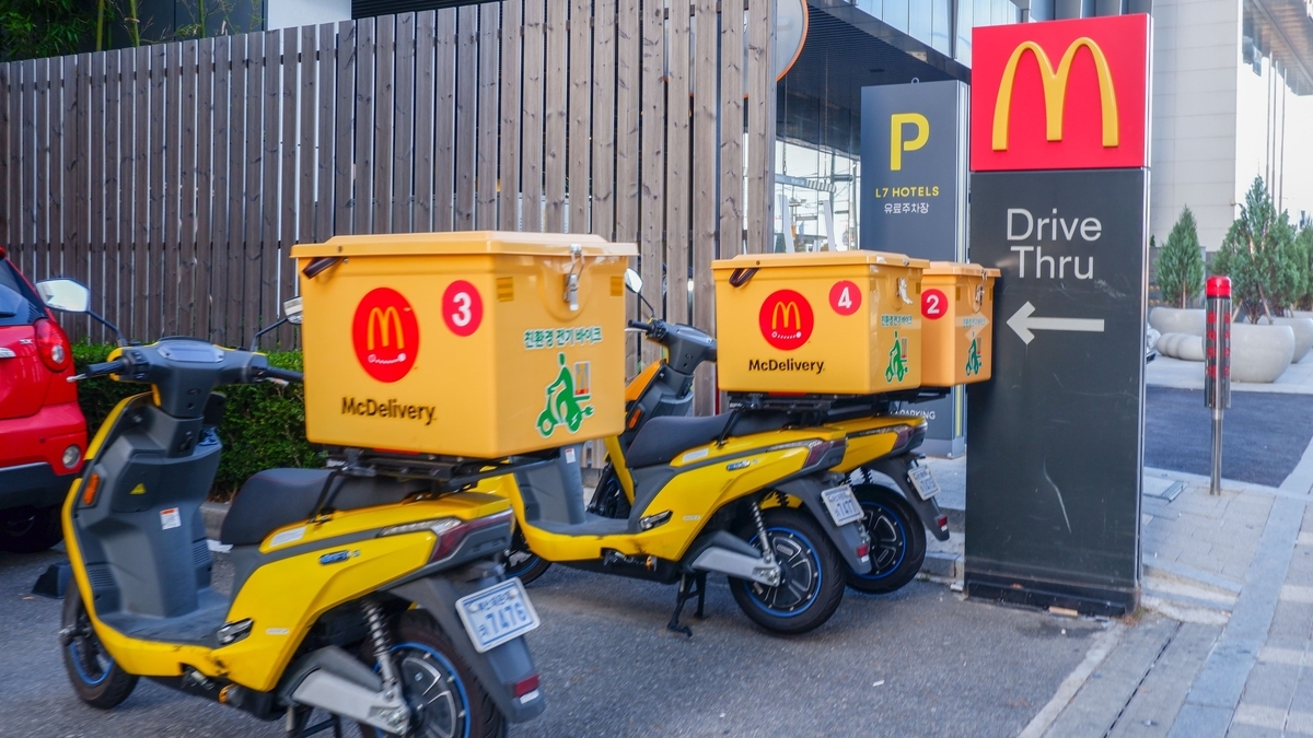 マクドナルドのデリバリー用ロゴが付いた黄色い電動バイクが、ドライブスルーの看板横に3台並んでいる。後部には大きな配送用ボックスが積載されている