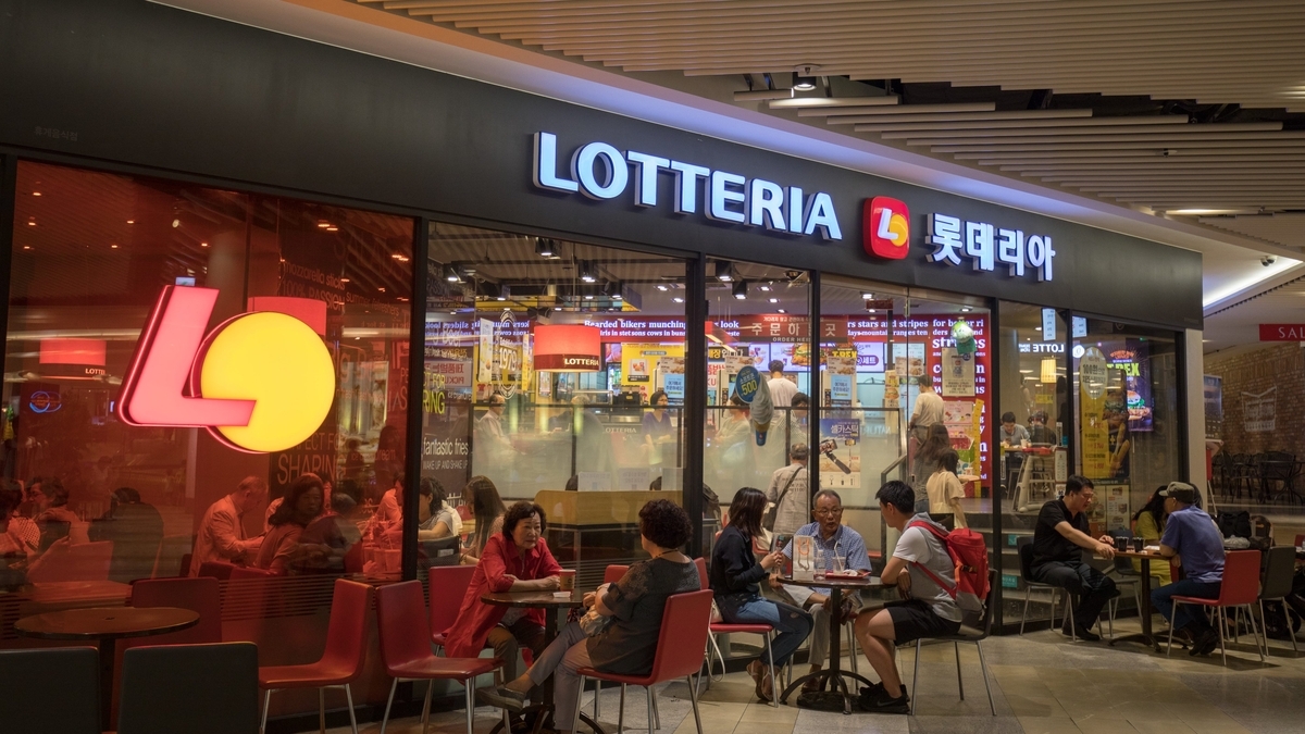 ロッテリア（LOTTERIA）の看板が光る、夜の店舗外観。ガラス張りの店内では、多くの人々がテーブル席に座って食事や会話を楽しんでいる