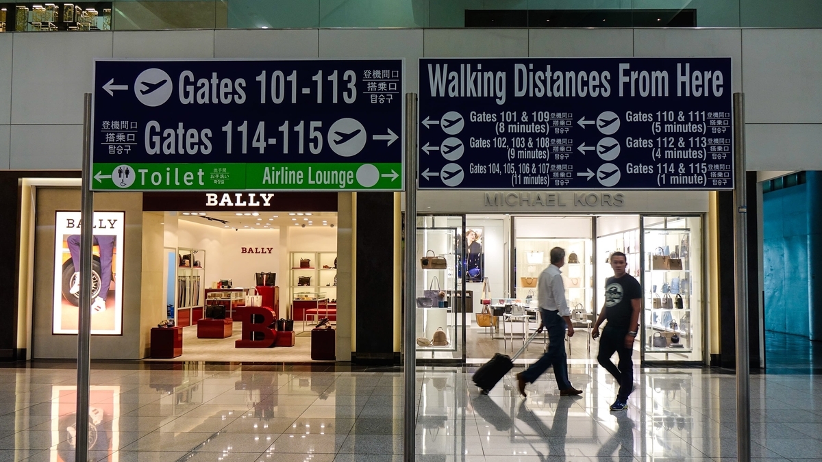 空港内にある案内板。左方向に搭乗口101-113番やトイレ、右方向に搭乗口114-115番や航空会社ラウンジを示している。下部にはBALLYとMICHAEL KORSの店舗が並んでいる