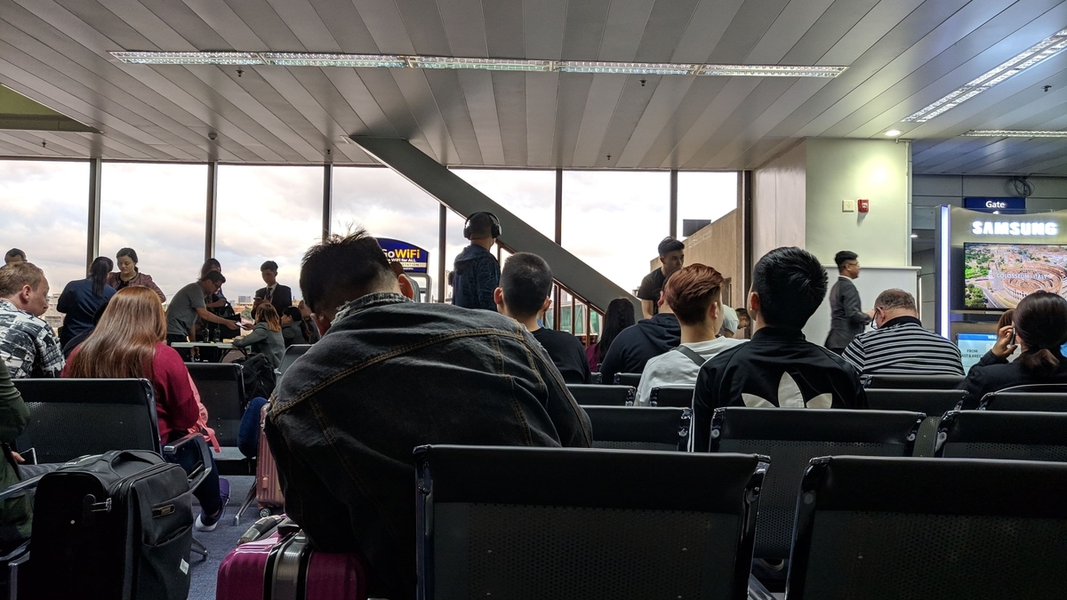 空港の搭乗待合室で椅子に座り出発を待つ人々。手前にはジーンズジャケットを着た男性の背中やピンク色のスーツケースが見える。窓の外は曇り空で、室内にはサムスンの広告ディスプレイが設置されている