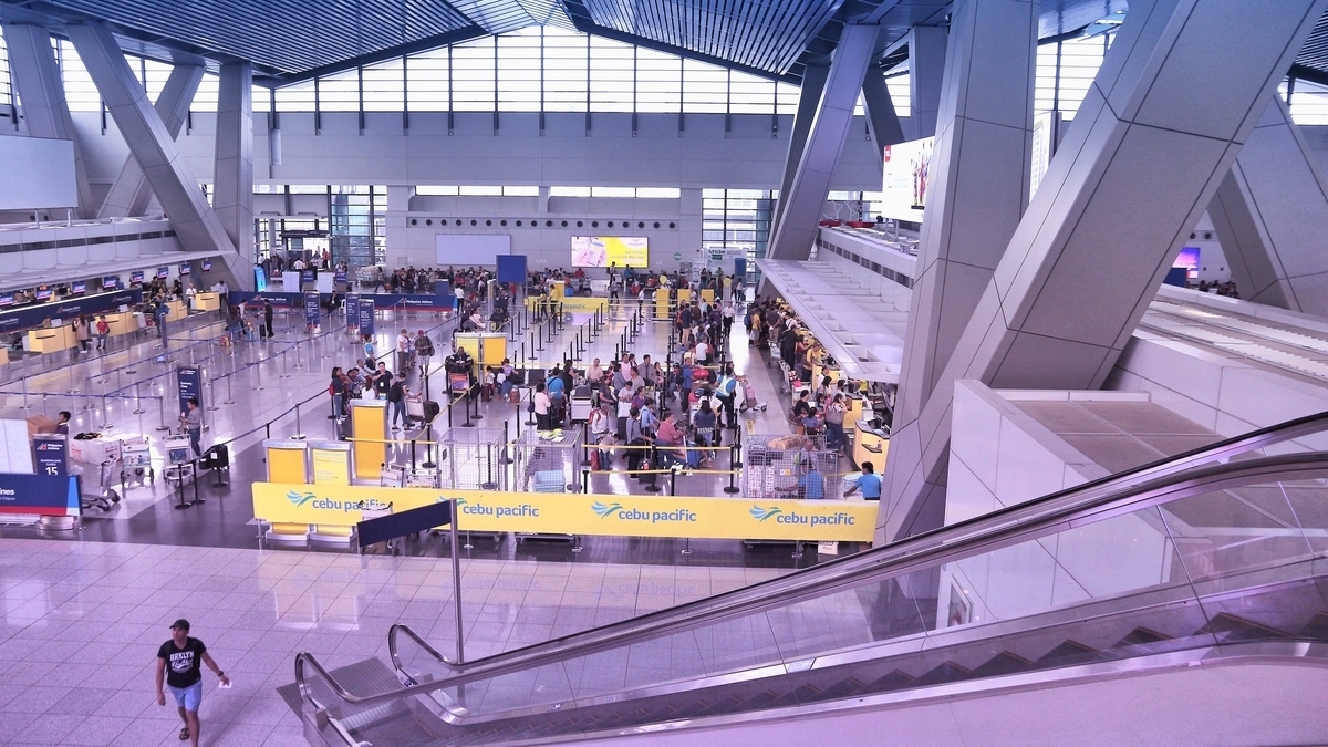 空港のチェックインカウンター。黄色いセブパシフィック航空の看板が置かれた搭乗手続きエリアに多くの乗客が並んでいる。吹き抜けの広い空間には巨大なV字型の柱が立ち、手前には下りエスカレーターがある