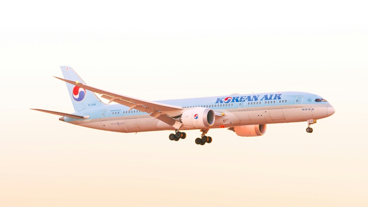 夕暮れ時の空を飛行する大韓航空のボーイング787-9型機。ライトブルーとシルバーの機体に紺色のロゴが描かれ、着陸装置を下ろした状態で水平に飛んでいる。背景は淡いオレンジ色のグラデーションに包まれている