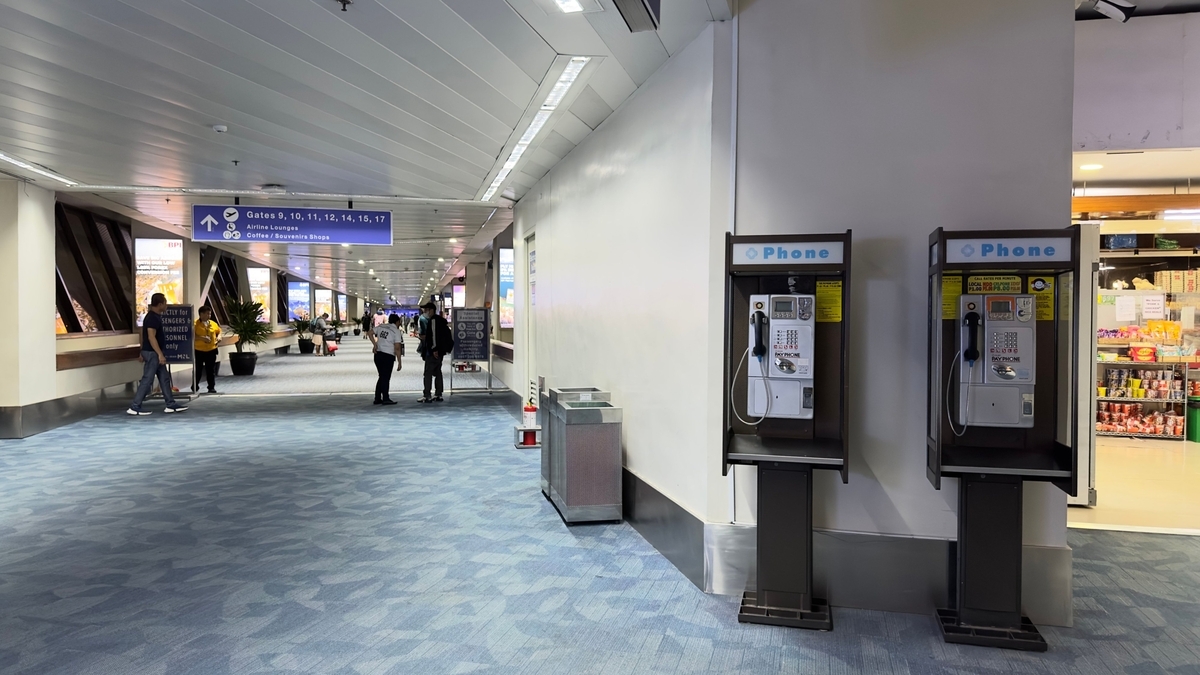 空港内の青いカーペットが敷かれた通路に設置された2台の公衆電話。奥には搭乗ゲート案内板や移動する人々の姿、右側には売店の棚が見える