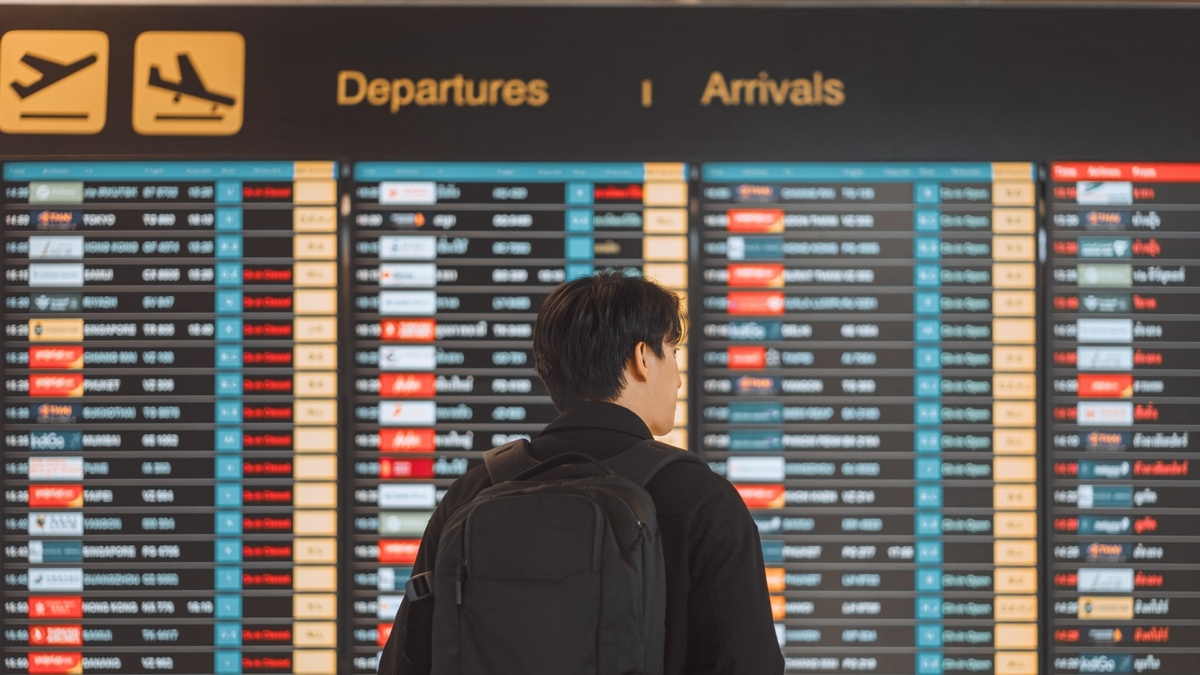 バックパックを背負った男性が、空港の大きな電光掲示板を見上げている後ろ姿。掲示板には出発と到着の便名や行き先が多数表示されている