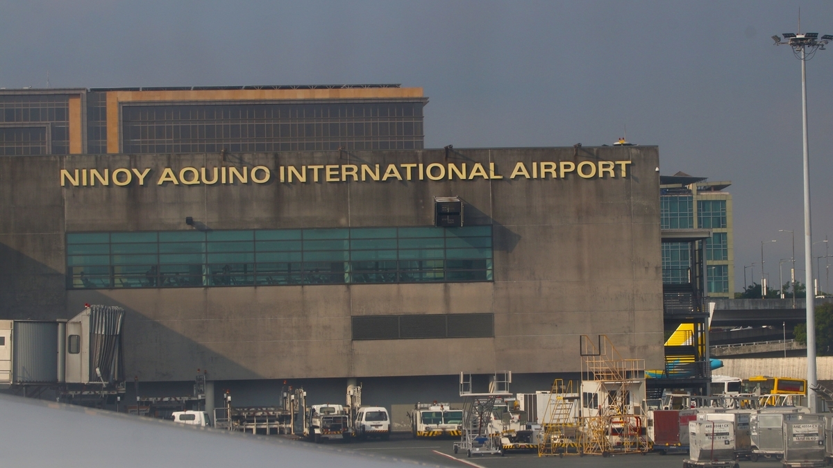 「NINOY AQUINO INTERNATIONAL AIRPORT」の文字が掲げられた空港の建物。手前の地上階には、白い作業車両や貨物用コンテナが並んでいる