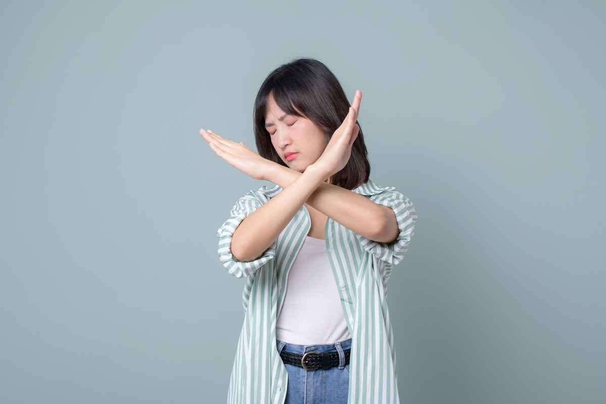 緑と白のストライプシャツを着た女性が、灰色の背景の前で、両腕を顔の前でクロスさせ、目を閉じて「NG」のポーズをとる様子