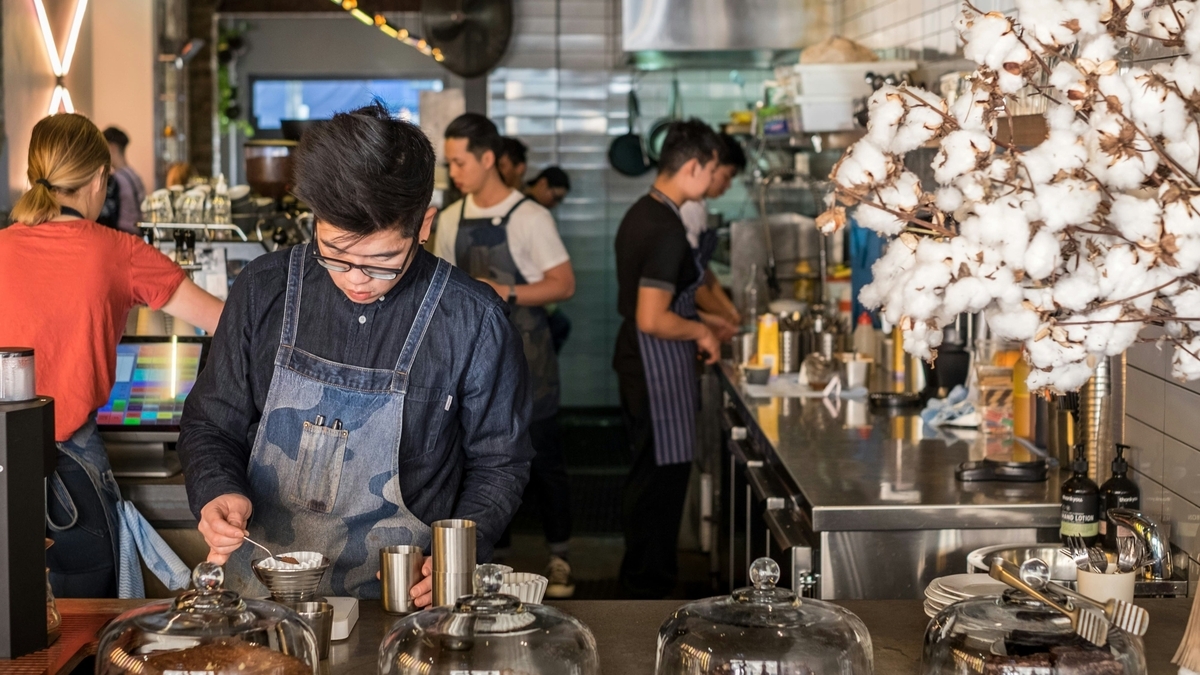 活気のあるカフェの厨房で、デニムのエプロンをつけた男性店員が真剣な表情でコーヒーを淹れている。カウンターにはケーキドームが並び、奥では他のスタッフが調理作業をしている