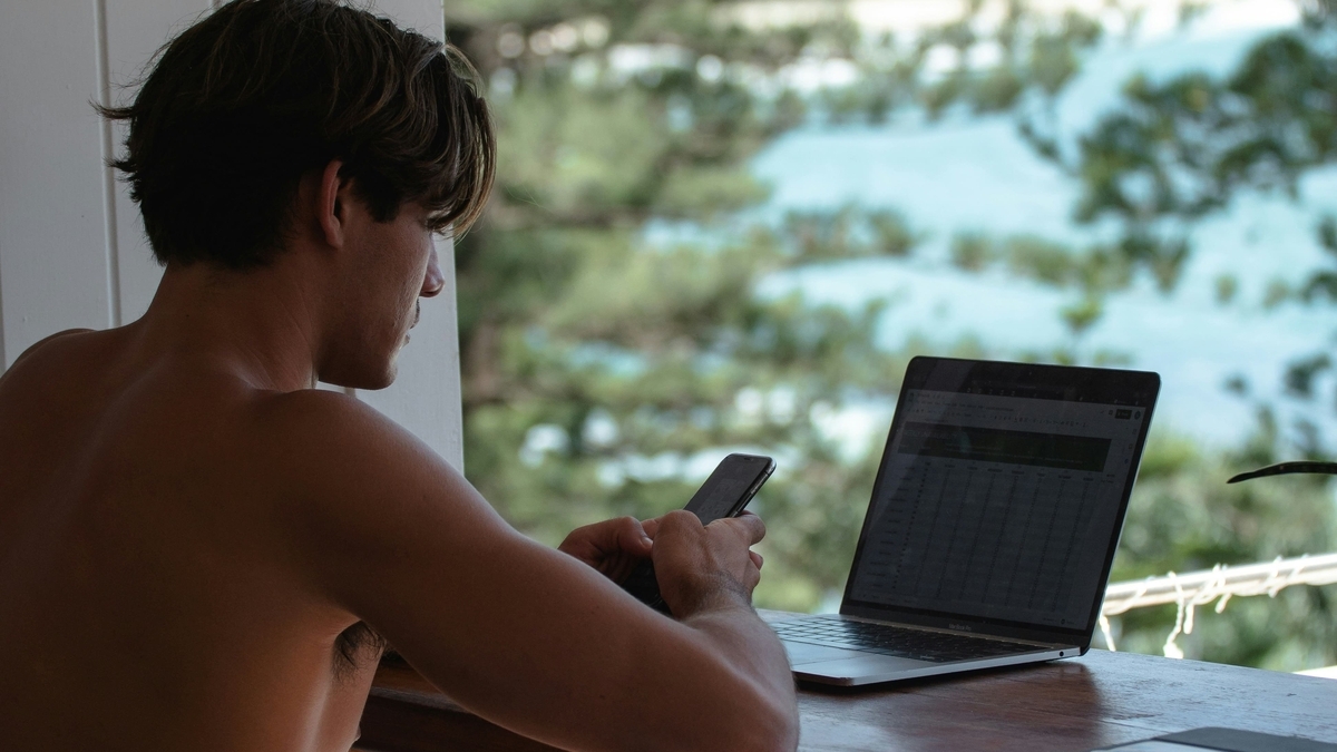 海が見える開放的なテラス席で、上半身裸の男性がノートパソコンを広げ、スマートフォンを操作している様子。木製のカウンターにはタブレット端末も置かれ、遠くには海岸線が広がっている