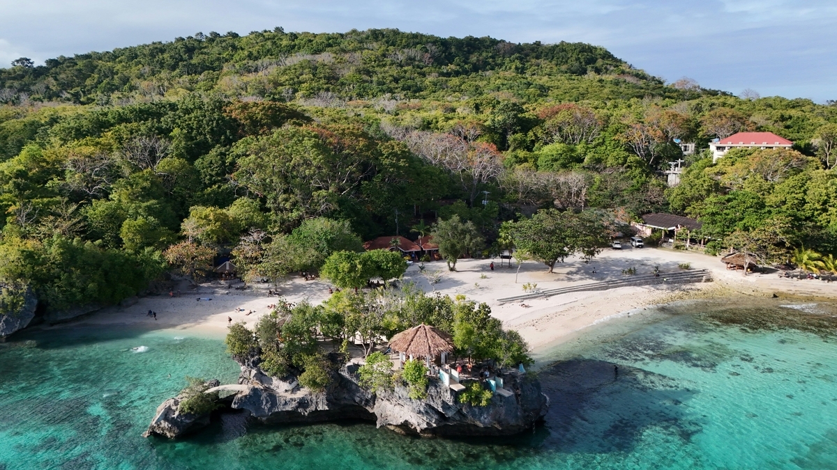 透明度の高いエメラルドグリーンの海に囲まれた、緑豊かな小島の風景。白い砂浜が広がる岸辺には茅葺き屋根の東屋が立ち、背後にはこんもりとした熱帯の木々が茂っています