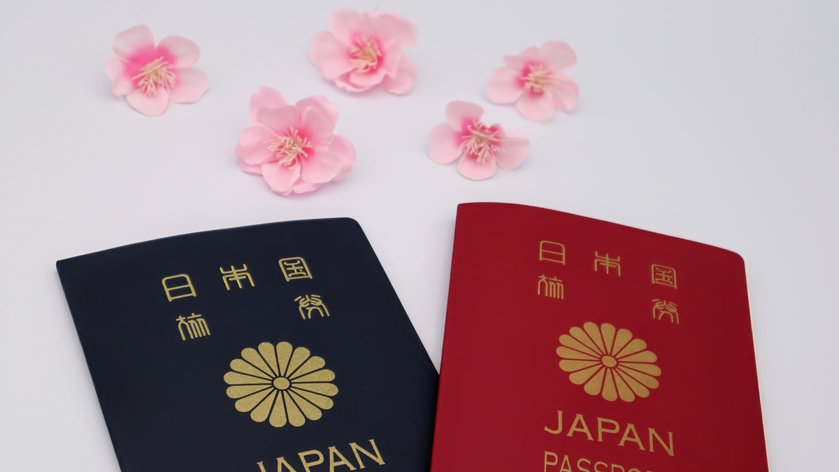 白を基調とした背景に並べられた、紺色と赤色の2冊の日本国パスポート。中央には十六一重表菊の紋章が金色で印字されており、上部には淡いピンク色の桜の花が5輪、彩りとして添えられています