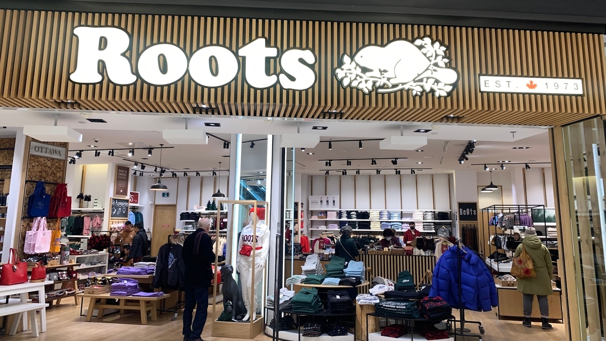 ショッピングモール内にあるRootsの店舗外観。木目調の壁にブランドロゴとビーバーのマークが掲げられ、店内には棚に整然と並べられた衣類や、マネキン、買い物をする客の姿が見える
