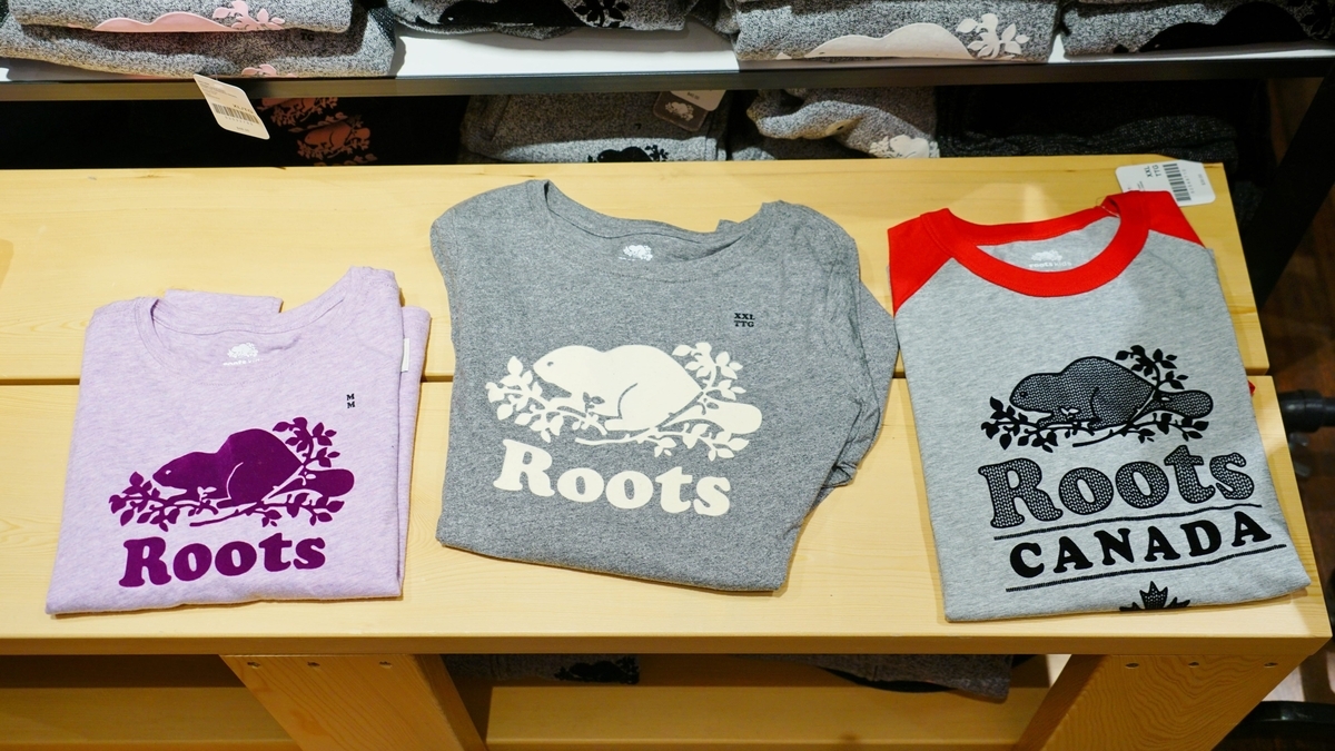 薄紫色の生地に濃い紫のロゴ、グレーの生地に白のロゴ、グレーに赤い袖のラグランTシャツの3枚が、木製の棚の上に並べて置かれている。いずれも中央にビーバーのイラストとRootsの文字がプリントされている